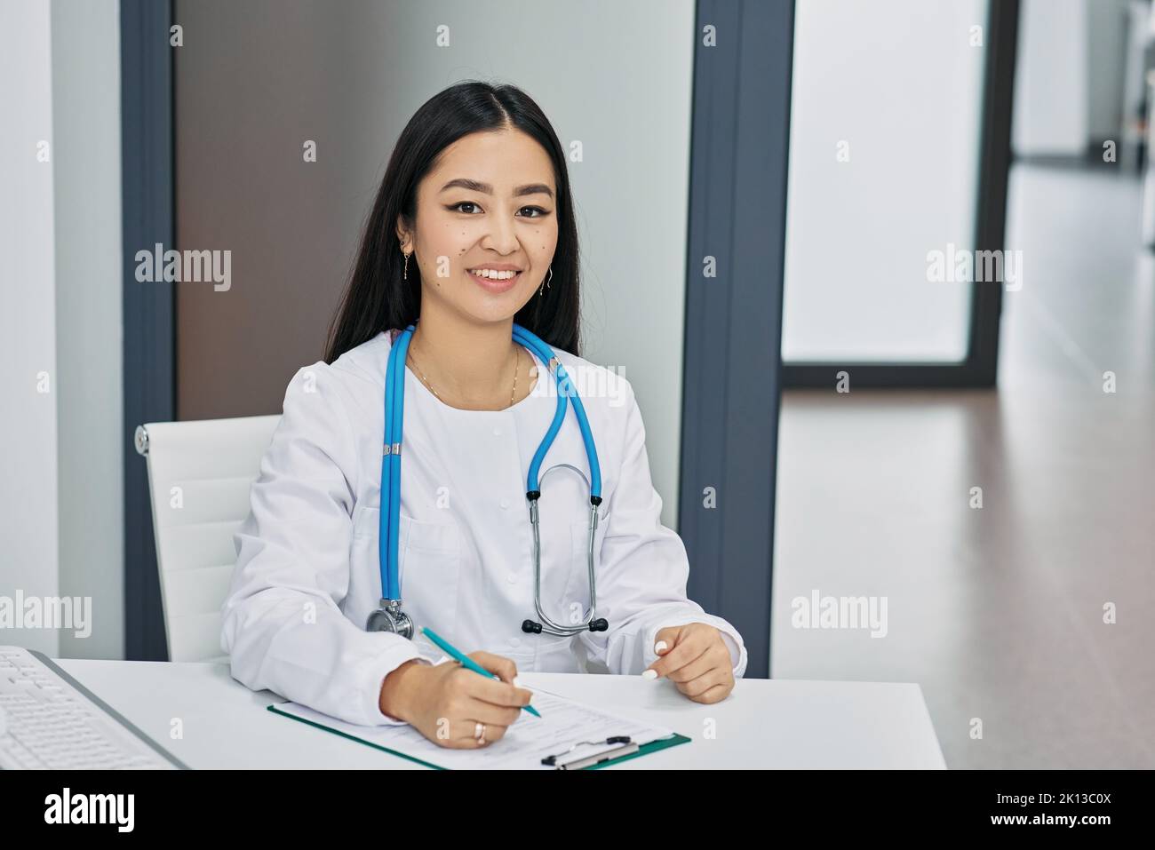 profession de médecin. Généraliste coréenne souriante assise sur son lieu de travail portant un uniforme médical Banque D'Images