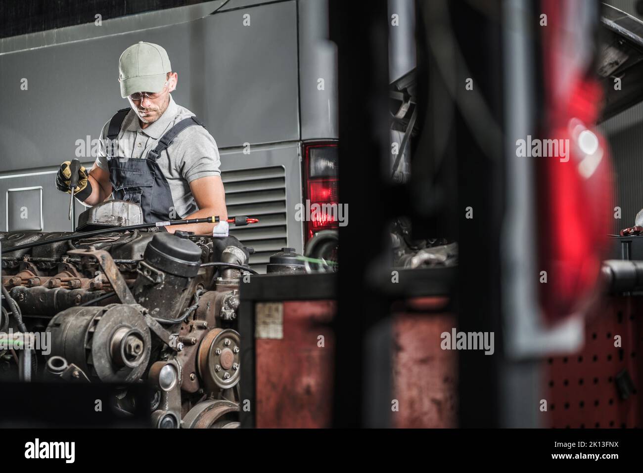 Mécanicien d'âge moyen de race blanche avec un tournevis dans sa main contrôle et réparation de gros moteur d'entraîneur dans son atelier de réparation de voiture. Entretien du véhicule Banque D'Images