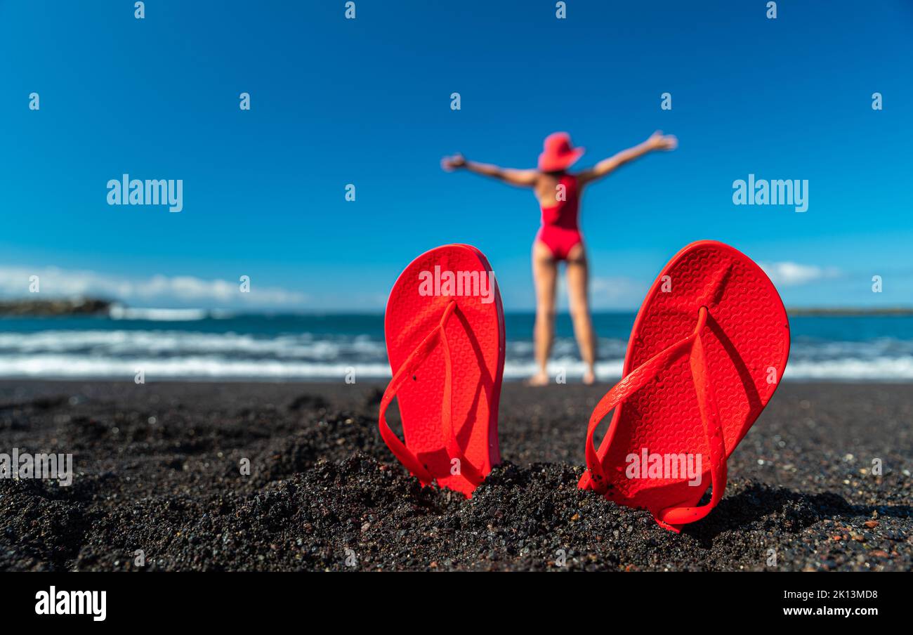 Tongs rouges dans le sable noir et silhouette de femme en maillot de bain rouge debout sur la plage de l'océan. Concept d'été. Banque D'Images