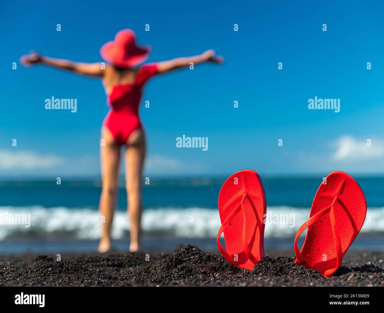 Tongs rouges dans le sable noir et silhouette de femme en maillot de bain rouge debout sur la plage de l'océan. Concept d'été. Banque D'Images