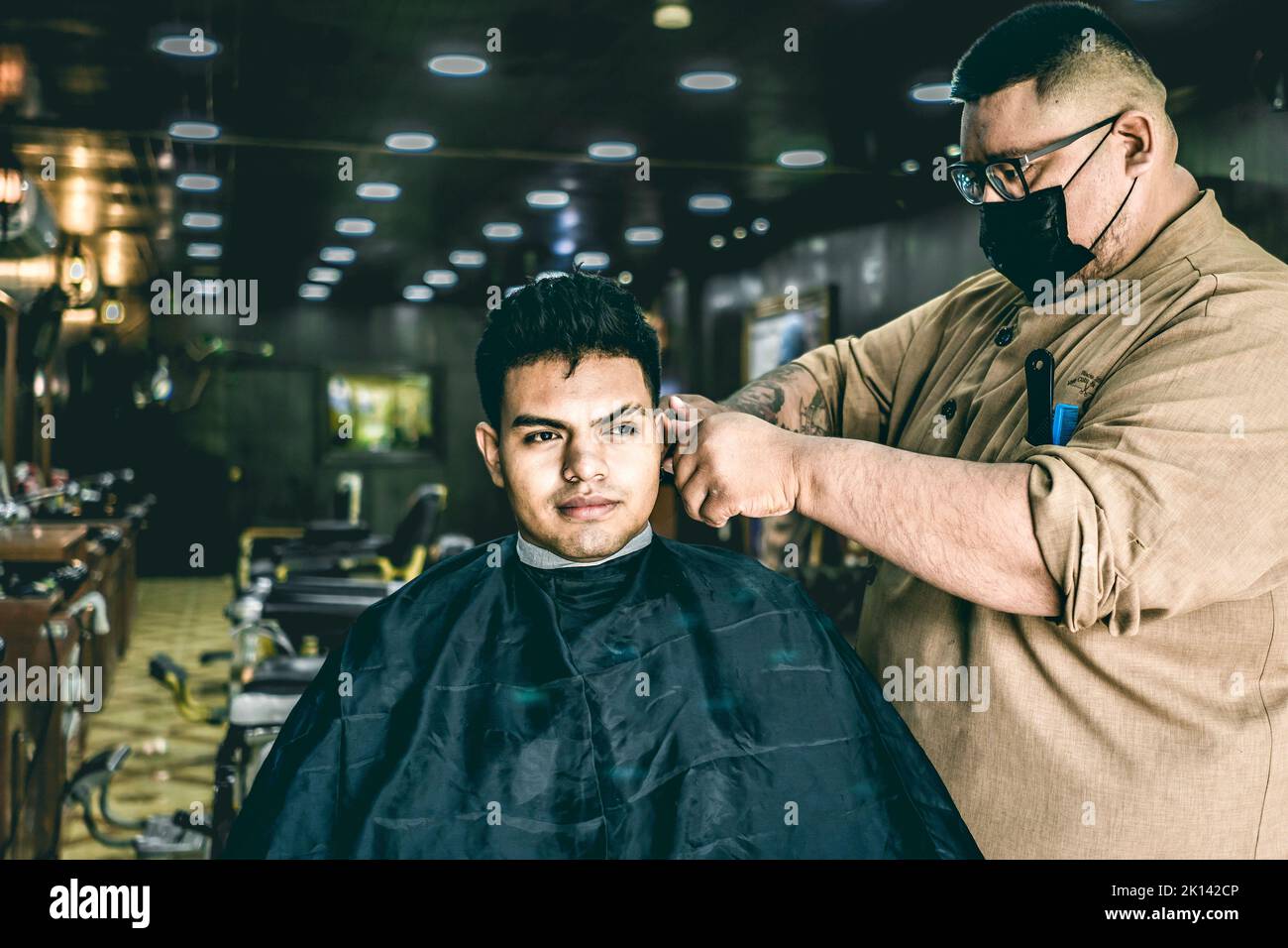 Coiffeur avec masque donnant une coupe de cheveux à l'homme Latino dans le salon de coiffure Banque D'Images