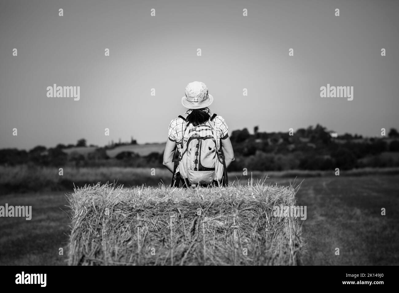 Photo de vue arrière en noir et blanc d'une femme randonneuse avec sac à dos et chapeau de soleil d'été, assise isolée sur une balle de foin regardant vers la campagne ouverte du Royaume-Uni. Banque D'Images