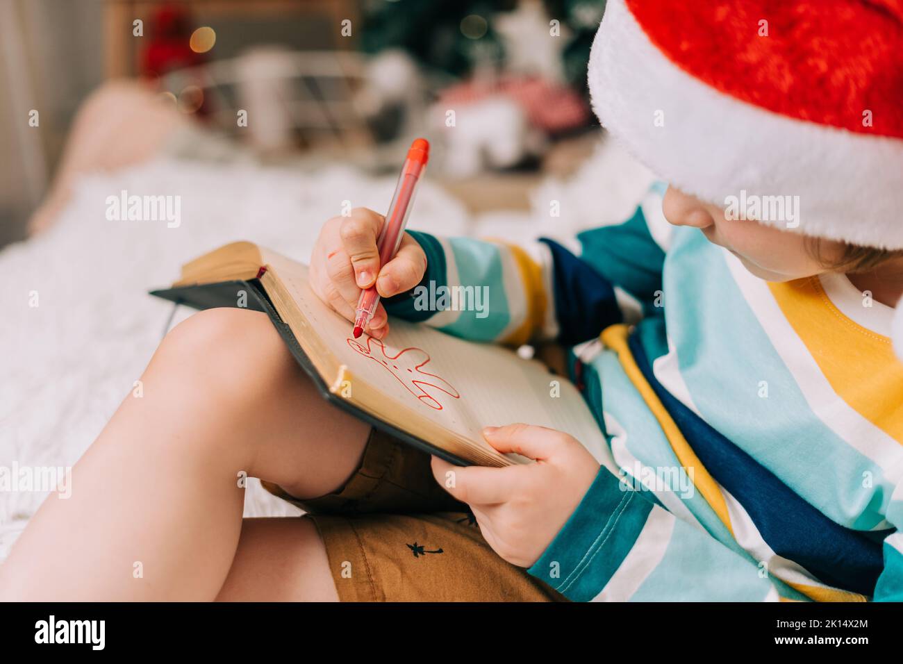 Enfant garçon dans le chapeau de père Noël rouge écrivant la lettre et le dessin à cher père Noël à la maison.Liste de souhaits enfant.Rêves de cadeaux de Noël.Joyeux Noël et heureux Banque D'Images