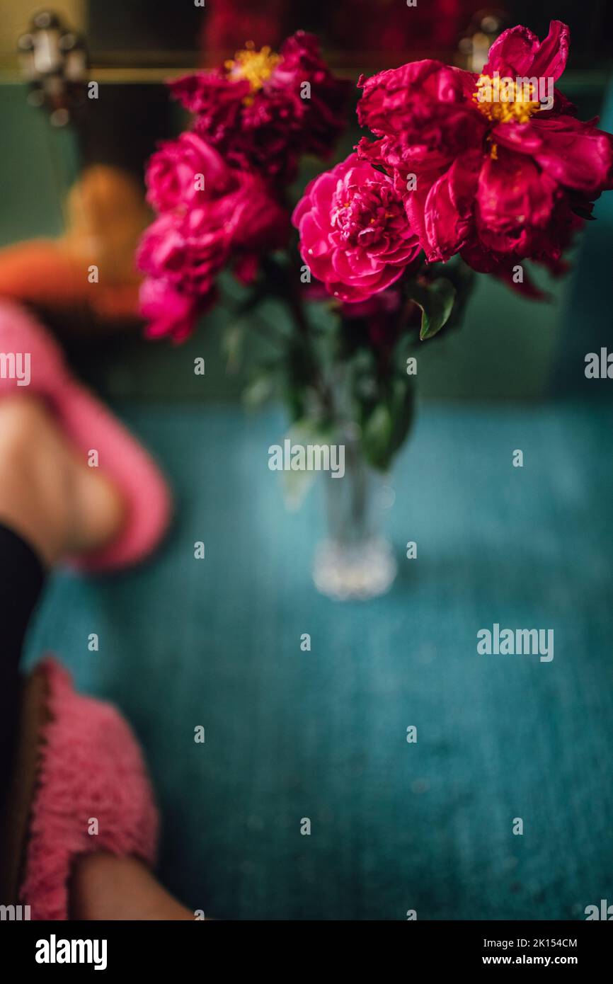 mourant de fleurs de pivoine rose magenta dans un vase en cristal sur un tapis de sarcelle avec une femme dans des chaussons roses flous Banque D'Images