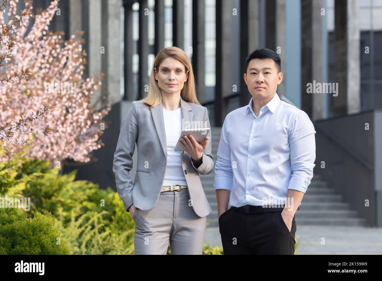 Portrait de deux hommes d'affaires interraciaux. Une jeune femme et un jeune homme asiatique se tiennent près du bureau en costume, regardant la caméra. Une femme tient une tablette. Un homme tient ses mains dans ses poches. Banque D'Images