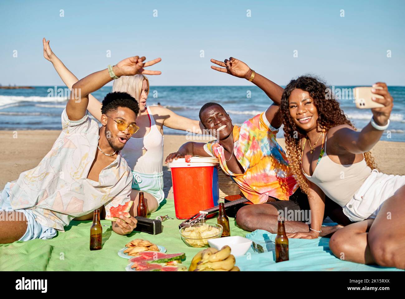Groupe de jeunes amis multiraciaux gais, hommes et femmes, vêtés de vêtements d'été souriant et montrant deux gestes de doigts tout en prenant le selfie sur smartphon Banque D'Images
