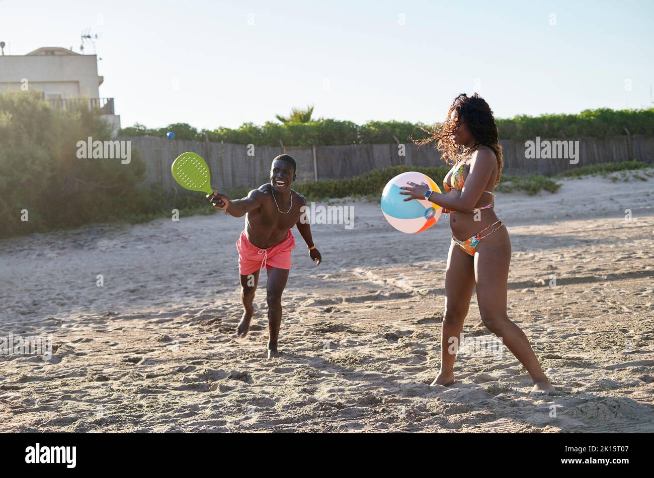 Joyeux Afro-américain jouant au tennis près d'une petite amie noire avec balle colorée tout en passant du temps sur la plage de sable par beau temps Banque D'Images