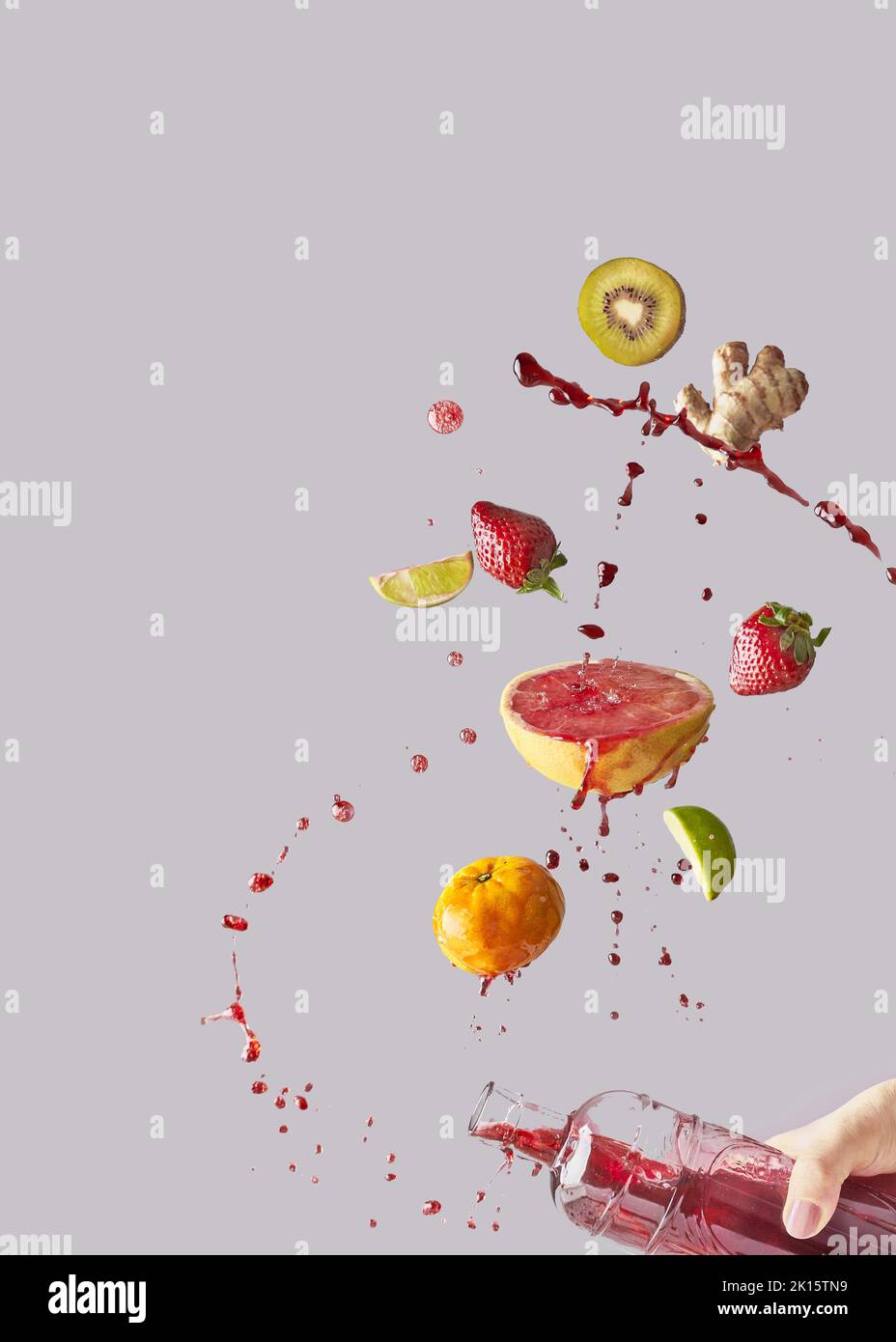 Bouquet de fruits frais divers et gingembre tombant dans une bouteille de verre étant maintenu par la personne anonyme de culture avec le jus sain sur fond gris Banque D'Images