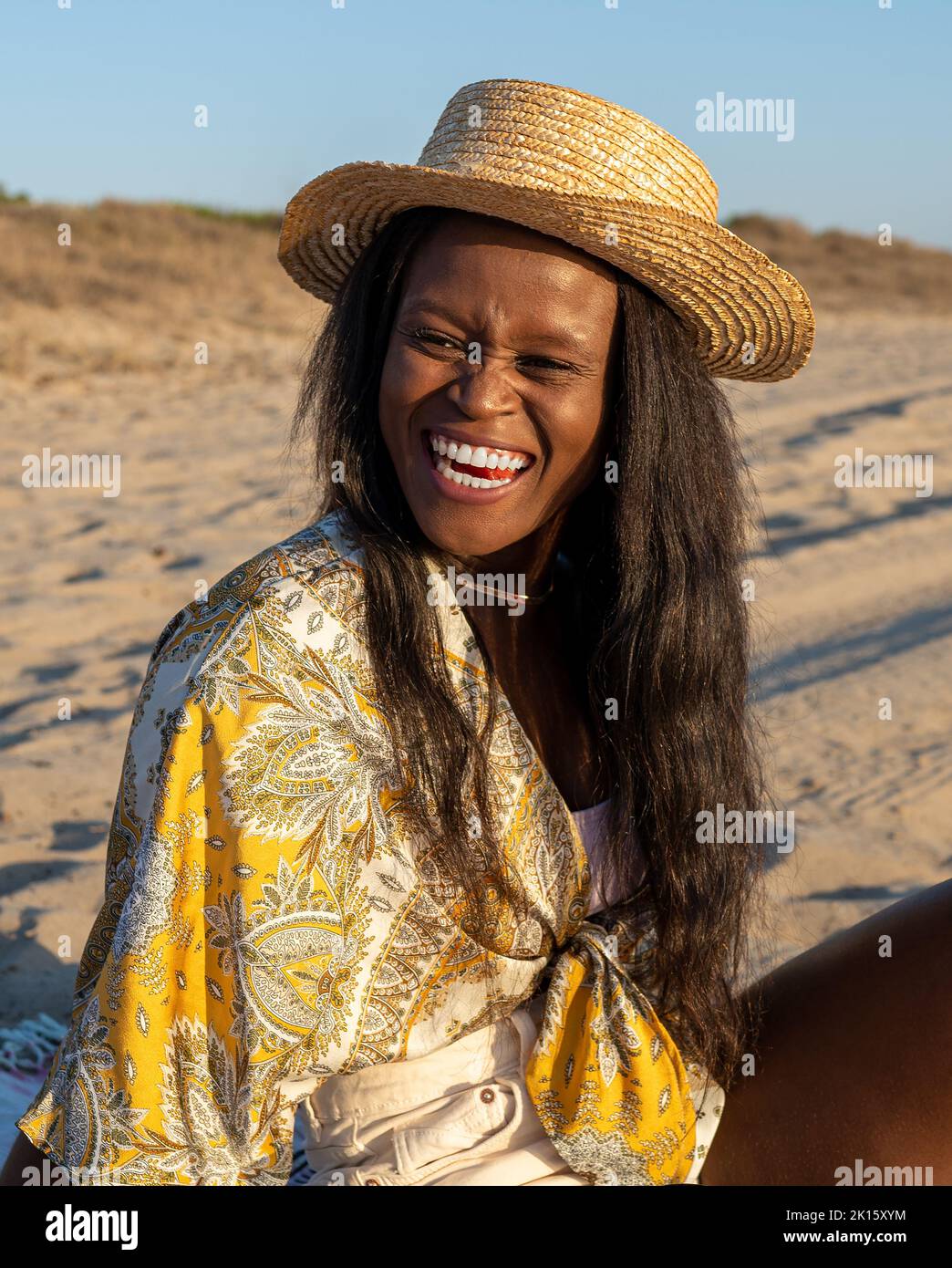 Joyeuse femme afro-américaine en vêtements de plage et chapeau de soleil regardant loin et riant à blague tout en passant une journée d'été ensoleillée sur la plage Banque D'Images