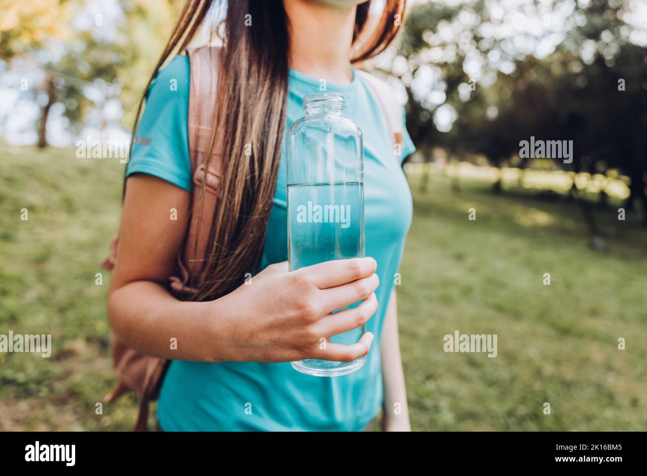 Fille méconnaissable portant un t-shirt turquoise et tenant une bouteille de verre avec de l'eau dans la nature. Ressources naturelles Banque D'Images