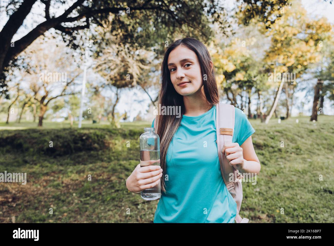 Jeune fille portant un t-shirt turquoise et un sac à dos rose, tenant une bouteille de verre avec de l'eau dans la nature. Ressources naturelles Banque D'Images