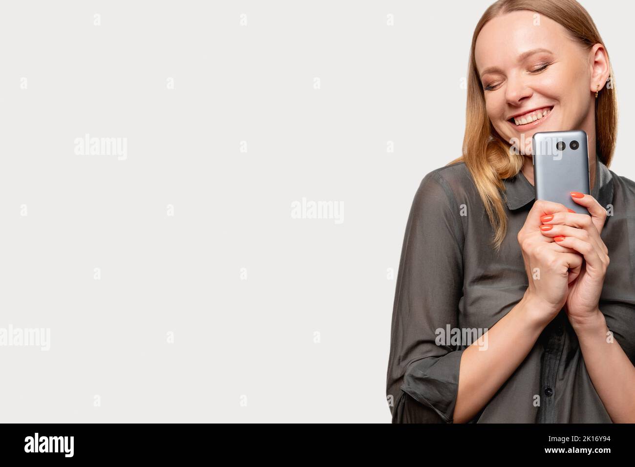 Technologie moderne. Femme heureuse. Un cadeau parfait. Appareil de gadget. Femme satisfaite en chemise grise tenant nouveau téléphone mobile souriant avec les yeux fermés isolé o Banque D'Images