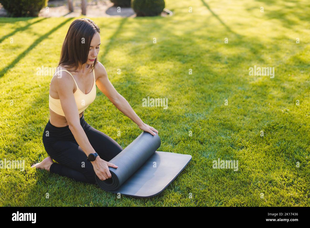 Caucasien mature femme assez mince en tenue sportive roulant son tapis de yoga après un entraînement dans le parc avec de l'herbe verte. Mode de vie sain, sport Banque D'Images