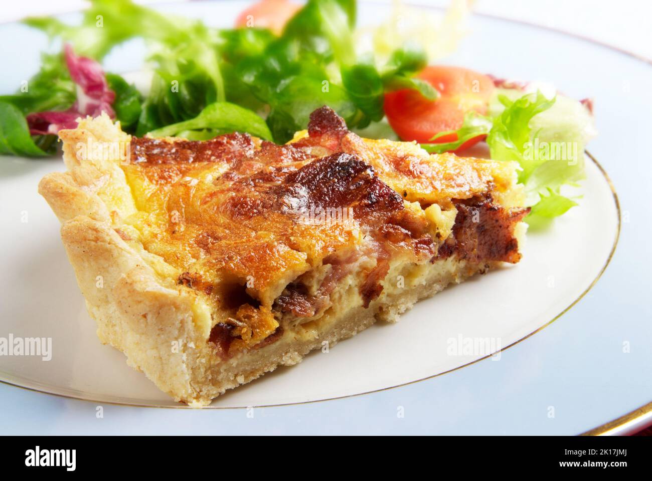 Une tranche de bacon et une quiche au fromage lorraine sur une assiette bleue avec une vinaigrette à la salade feuillue Banque D'Images