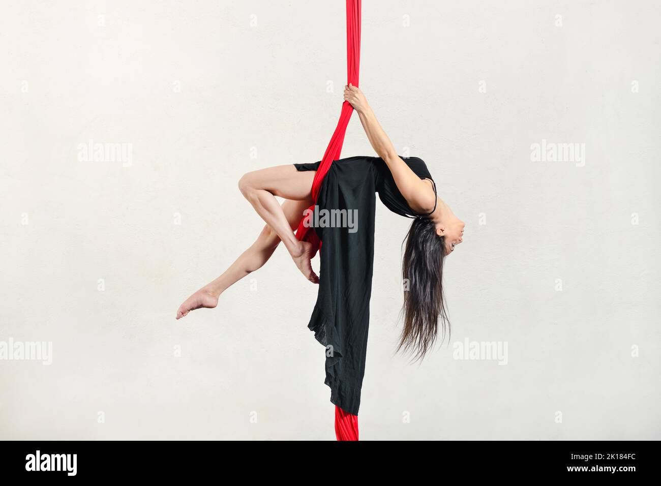 Vue latérale complète de la femme active aerialist effectuant un exercice d'oscillation sur des silks aériens sur fond blanc pendant l'entraînement en studio Banque D'Images