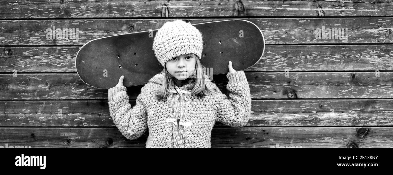 Bannière ou affiche supérieure horizontale avec portrait d'une fille skateboarder en laine vêtements d'hiver avec planche à roulettes sur ses épaules regardant l'appareil photo sur bois Banque D'Images