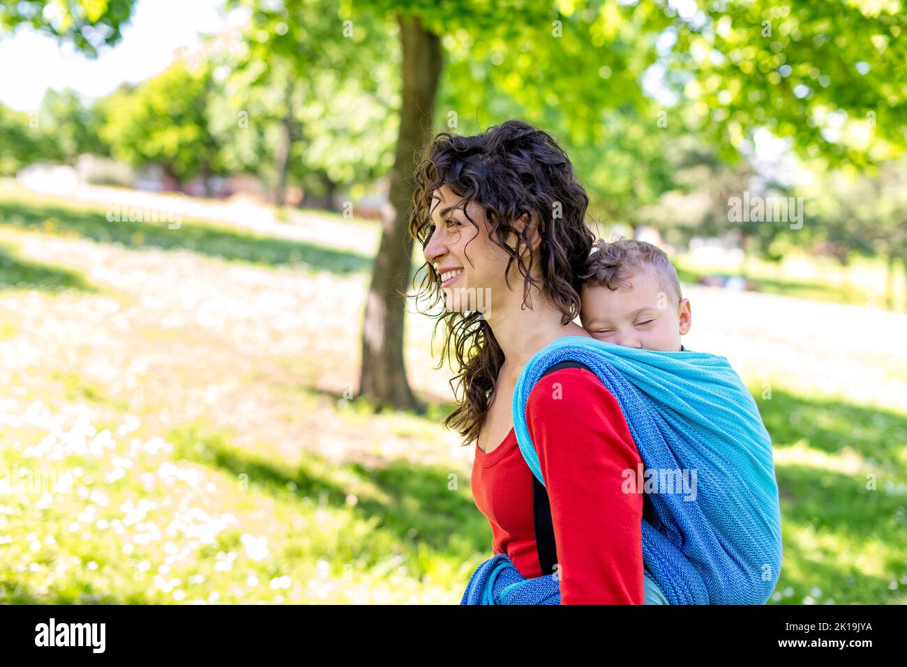 portrait rapproché d'une jeune maman heureuse porte un enfant sur son dos attaché avec un morceau de tissu traditionnel coloré dans un jardin de parc public. mère Banque D'Images