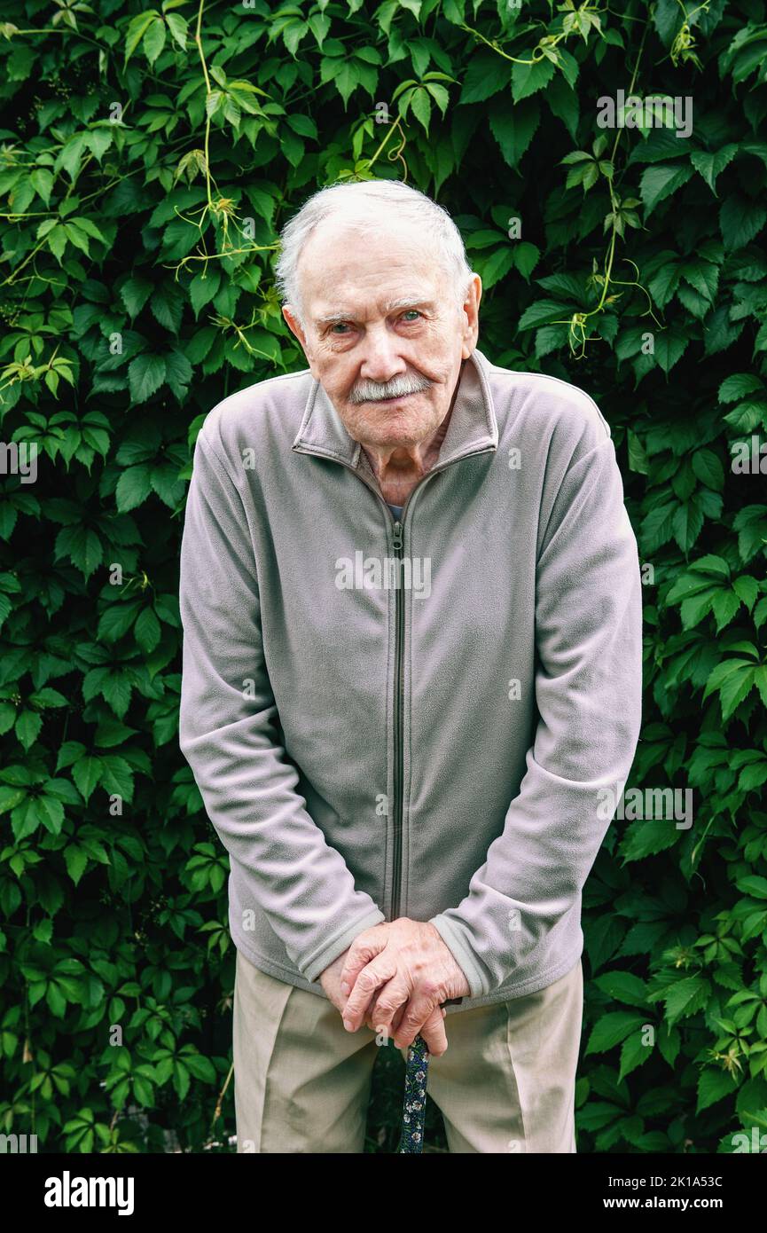 Un vieil homme de 88 ans avec une canne se dresse dans la rue devant un mur vert. Portrait d'un vieil homme aux cheveux gris avec une canne. Banque D'Images