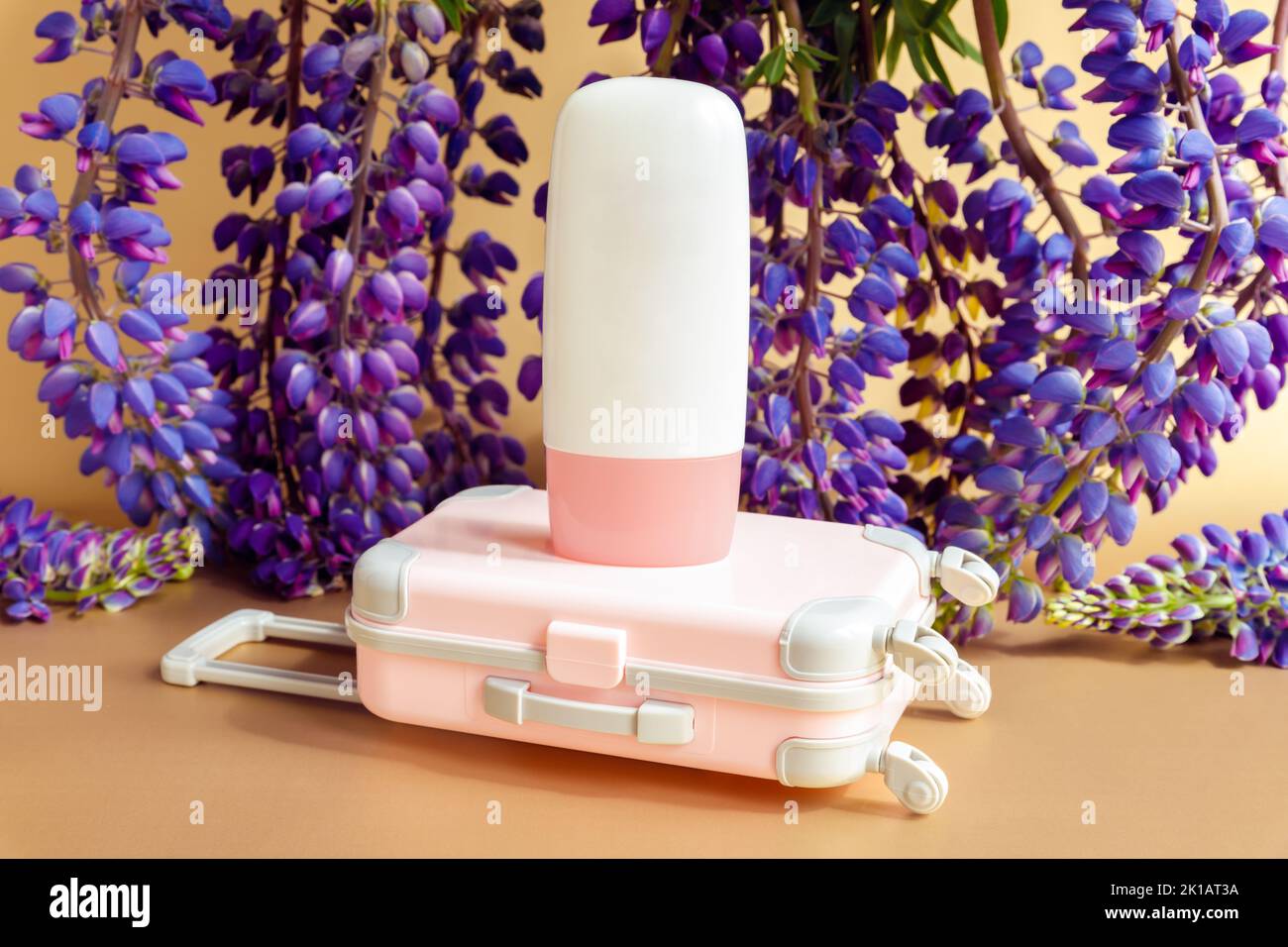 Bouteille de cosmétique Mockup blanche avec bouchon à vis rose, petite valise de voyage rose jouet et fleurs lupin pourpres beaucoup sur fond marron. Maquette, fro Banque D'Images