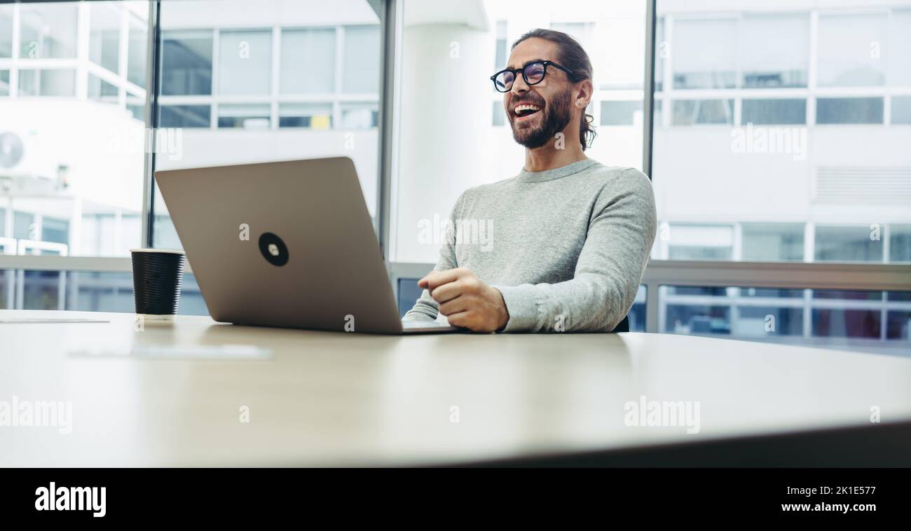 Un jeune homme d'affaires heureux riant avec joie tout en travaillant avec un ordinateur portable. Entrepreneur prospère travaillant à distance dans un espace de travail conjoint. Banque D'Images