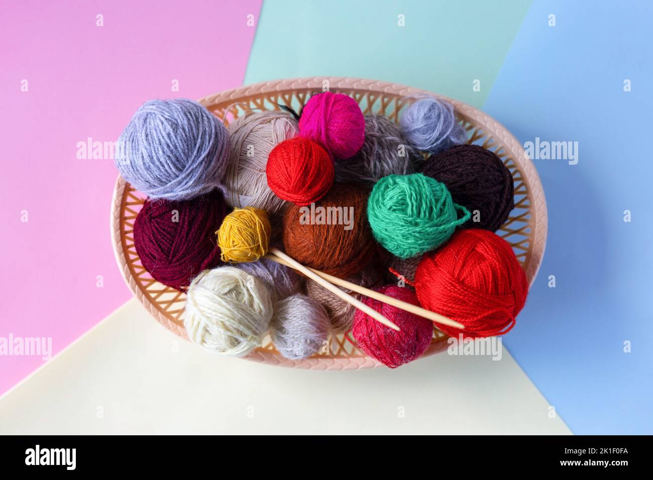 Rouge, jaune, blanc, violet, marron, boules vertes de fil dans un panier beige avec aiguilles à tricoter en bambou sur un fond multicolore. Banque D'Images