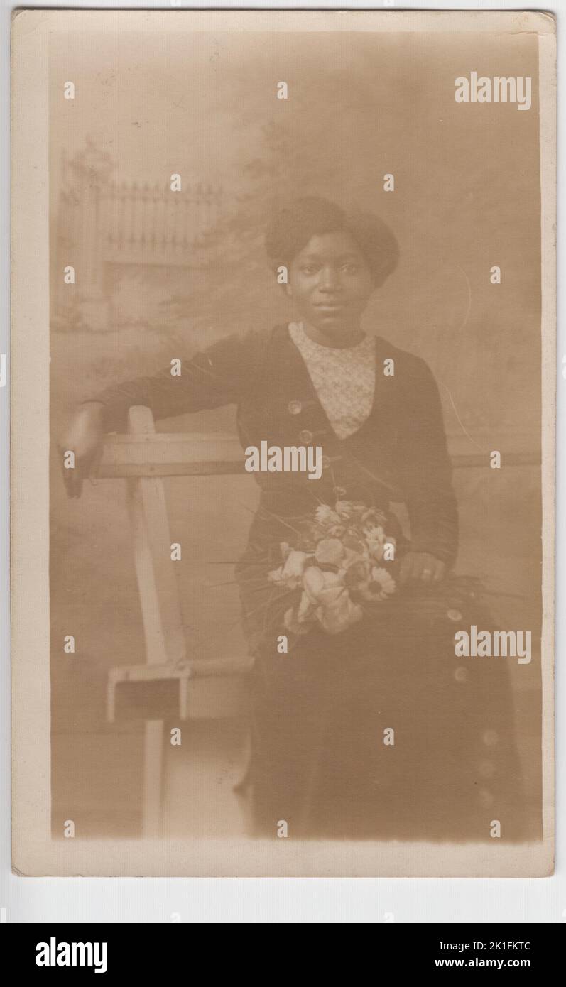 Portrait photographique d'une jeune femme britannique noire au début du 20th siècle. Elle est élégamment habillée et tendance et a été photographiée assis sur un banc dans un studio de photographe, tenant un bouquet de fleurs. L'image a été envoyée comme carte postale par Lily (la personne sur la photo) à Miss M Reade de Manor Farm, Balking, Berkshire, en janvier 1914 Banque D'Images