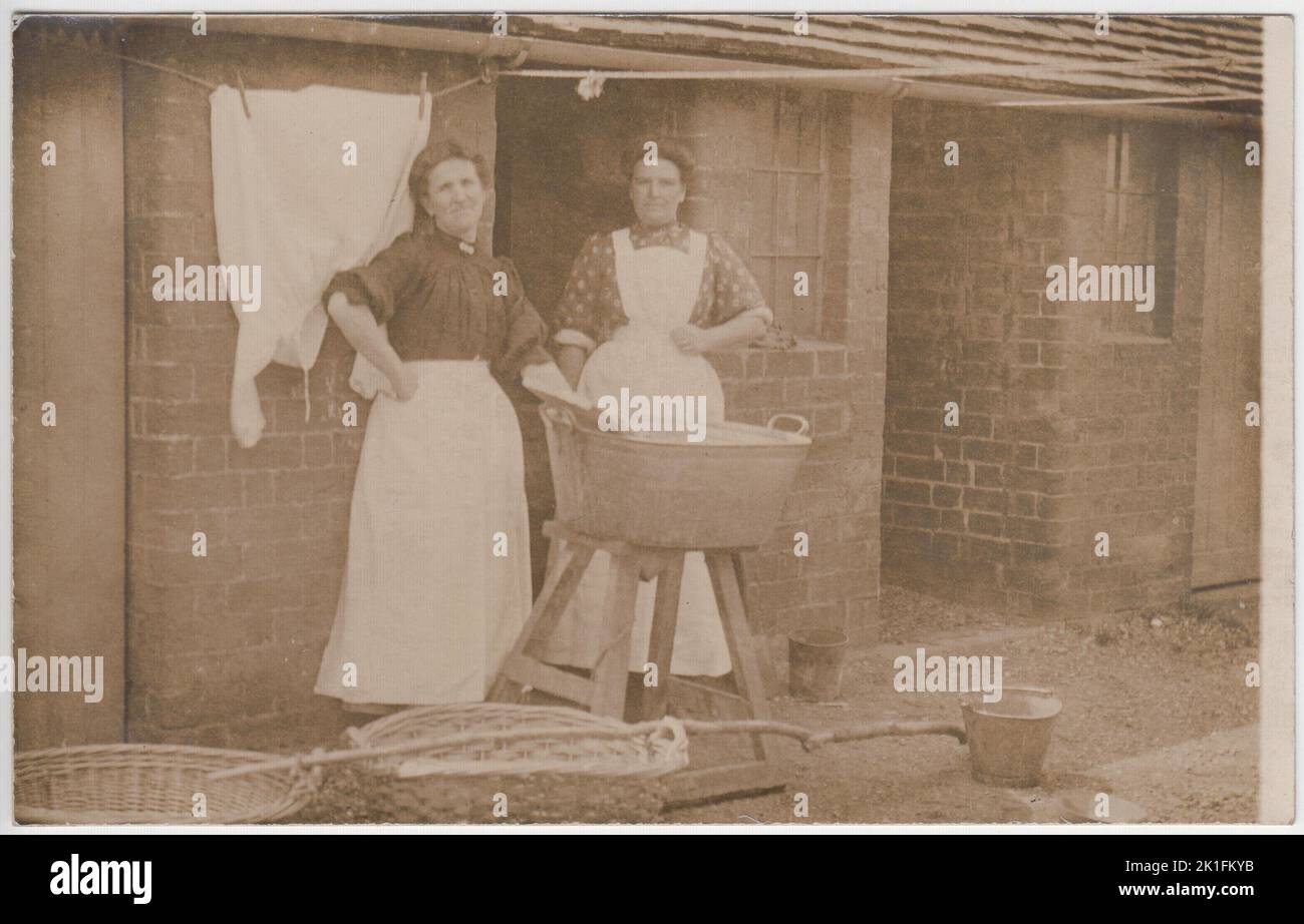 Journée de lavage dans le Bedfordshire d'Edwardian. Photo du début du 20th siècle de deux femmes vêtues de tabliers, debout près d'une grande baignoire de lavage qui a été équilibrée sur un tabouret. Deux paniers de linge en osier, un long bâton de transport et quelques seaux en métal sont sur le sol en face d'eux. Une chemise fraîchement lavée est accrochée à la ligne de lavage derrière elle. La photo porte le nom de deux photographes au dos - Ussher du 24, rue Battenberg, Bedford, et Lennox Gordon de Woburn Sands. Banque D'Images