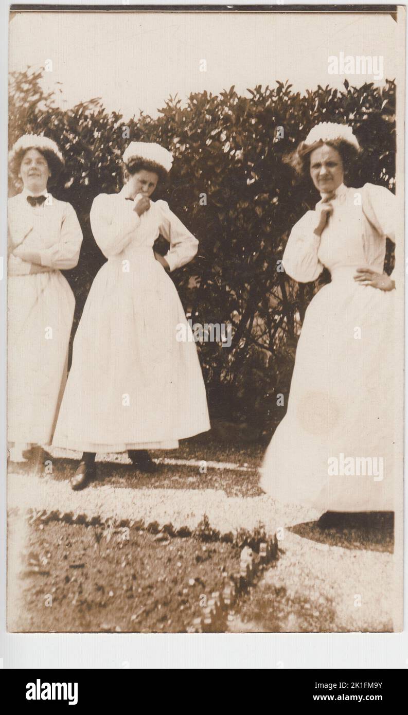 Portrait informel de trois serviteurs jouant autour de la caméra, début 20th siècle. Les femmes sont vêtues de leur uniforme (avec des chapeaux) et sont dans un jardin. Deux femmes sont debout avec une main sur leur hanche, et l'autre tenue de façon cyclique contre leur visage. La troisième femme de ménage rit dans le coin de la photo Banque D'Images