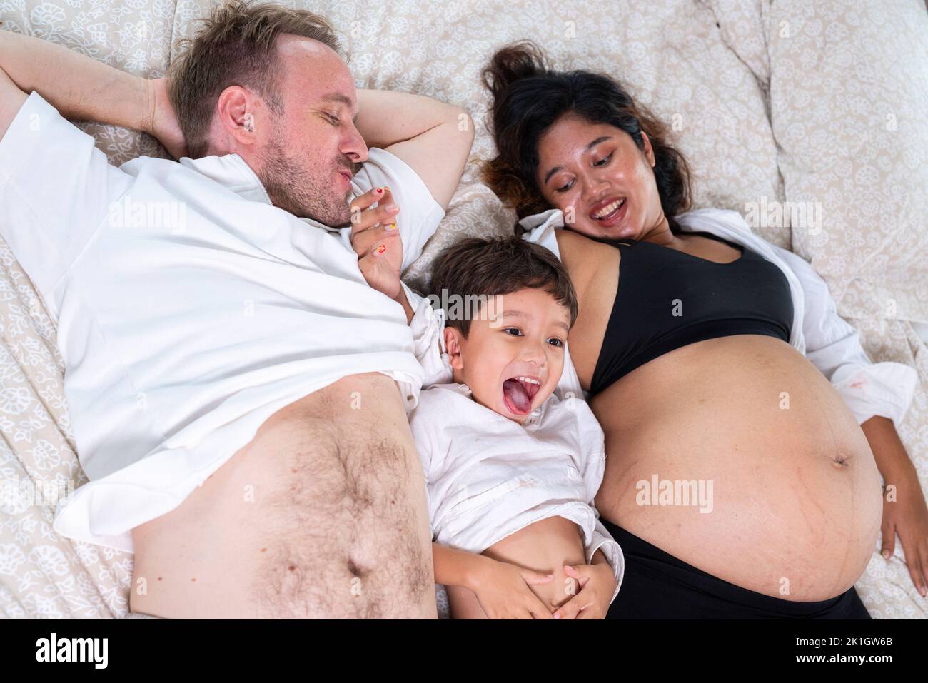 Photo de jeune couple gai positif souriant femme enceinte et mari avec leur fils se détendant à la maison Banque D'Images