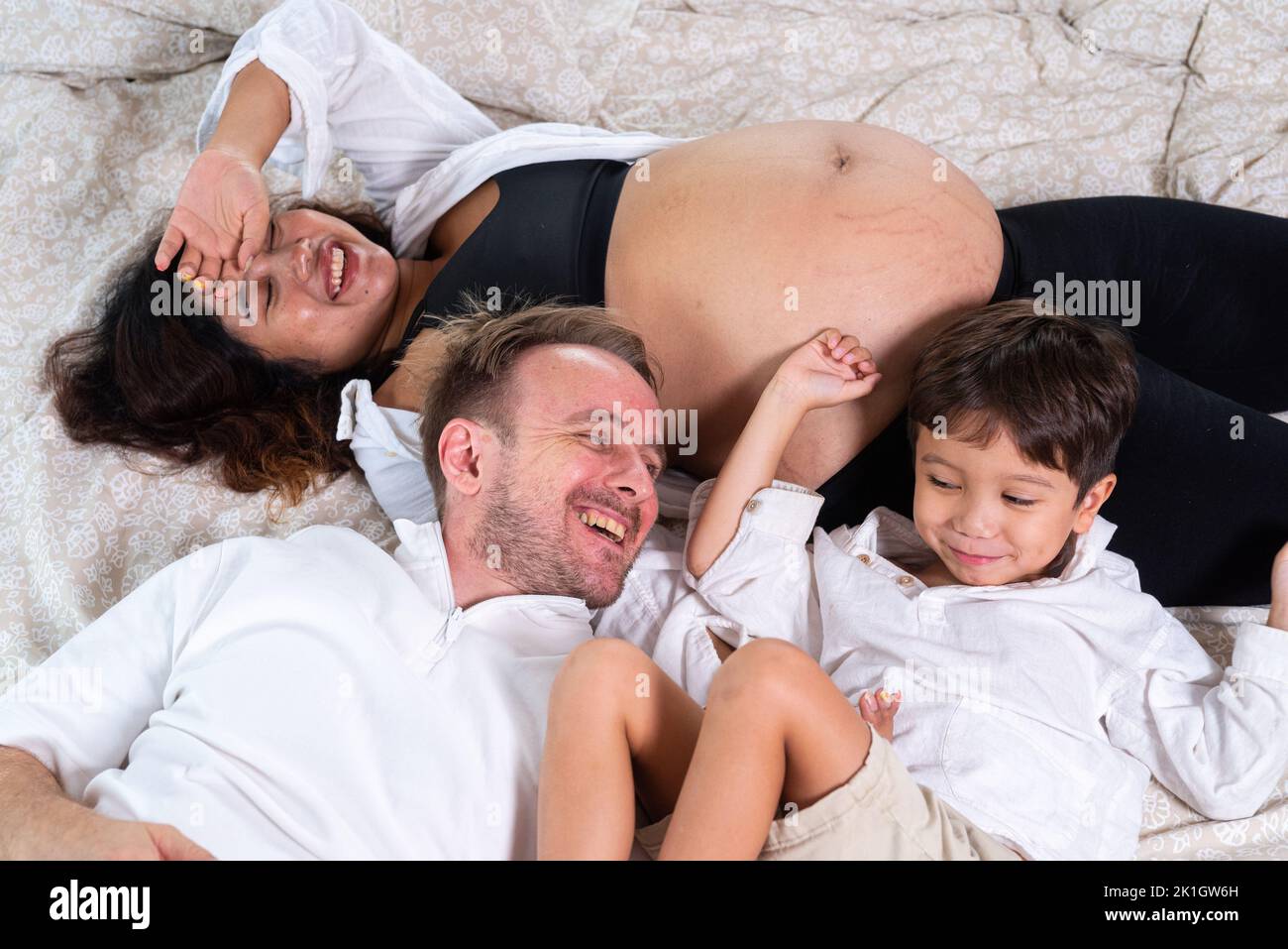 Photo de jeune couple gai positif souriant femme enceinte et mari avec leur fils se détendant à la maison Banque D'Images