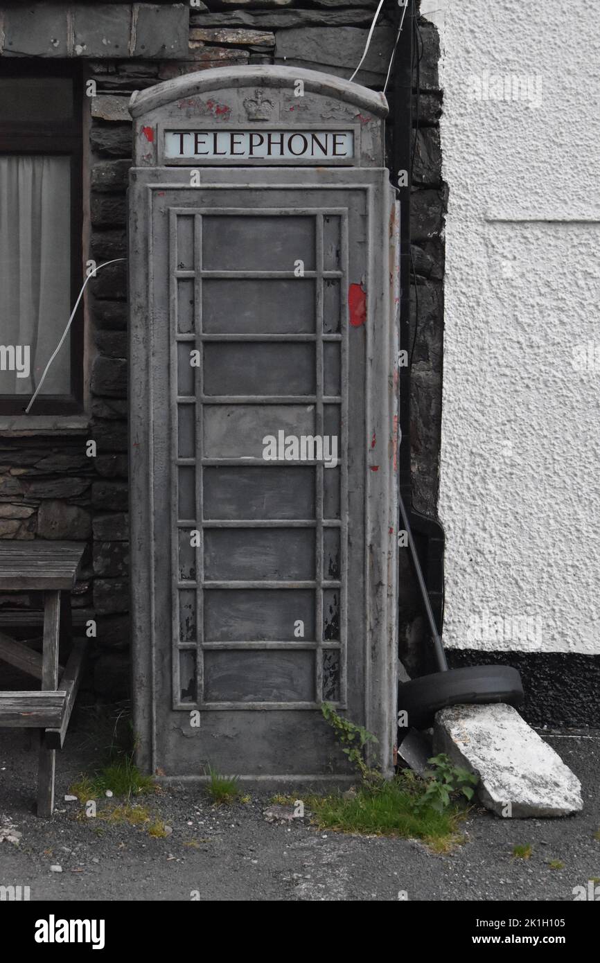 Une boîte téléphonique désutilisée à l'extérieur de Kirkstone Pass Inn, Ambleside, Lake District, Royaume-Uni. Gris, avec des éclats de peinture rouge et de mauvaises herbes vertes. Banque D'Images