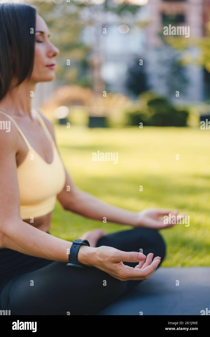 Vue latérale d'une femme en vêtements de sport pratiquant la méditation dans un parc urbain assis en position lotus sur son tapis de yoga. Femme de fitness. Un mode de vie sain Banque D'Images