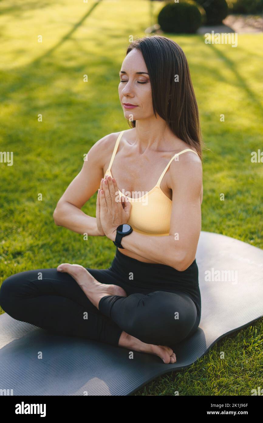 Brunette femme caucasienne mûre méditant dans le parc faisant du yoga, des sports, assis un tapis de yoga. Prendre soin de son corps et de sa santé mentale. Fitness Banque D'Images