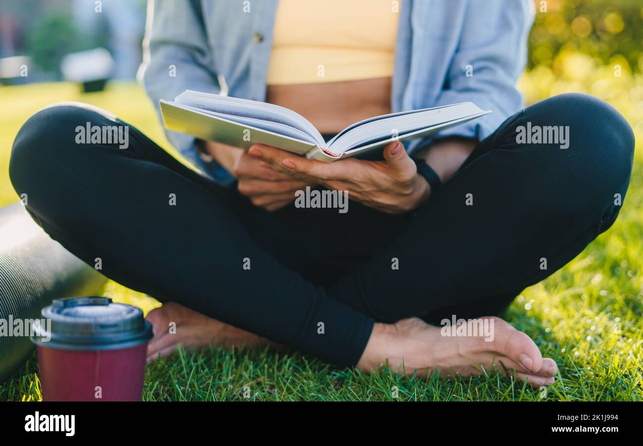 Mains d'une femme vêtue de vêtements de sport tenant un livre de culture générale, lisant après avoir pratiqué le yoga du matin dans le parc. Passe-temps de lecture. Extérieur Banque D'Images