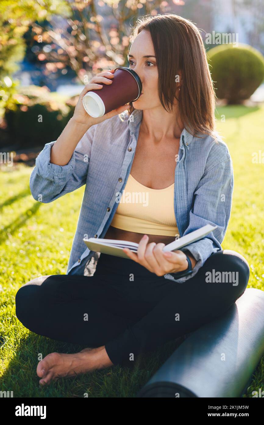 Portrait d'une femme caucasienne souriante buvant un café du matin, lisant un livre de texte assis près du tapis de yoga sur le terrain d'herbe. Style de vie de l'éducation en plein air Banque D'Images