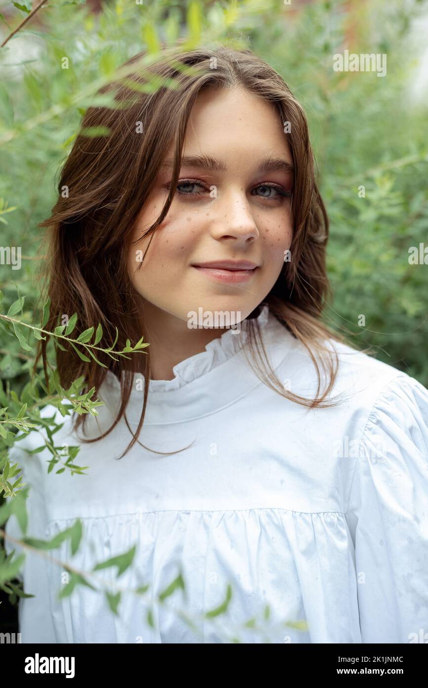 Portrait gros plan de jolie jeune fille en robe blanche avec plaisir debout parmi les branches de buissons avec des feuilles vertes, fond de la nature Banque D'Images