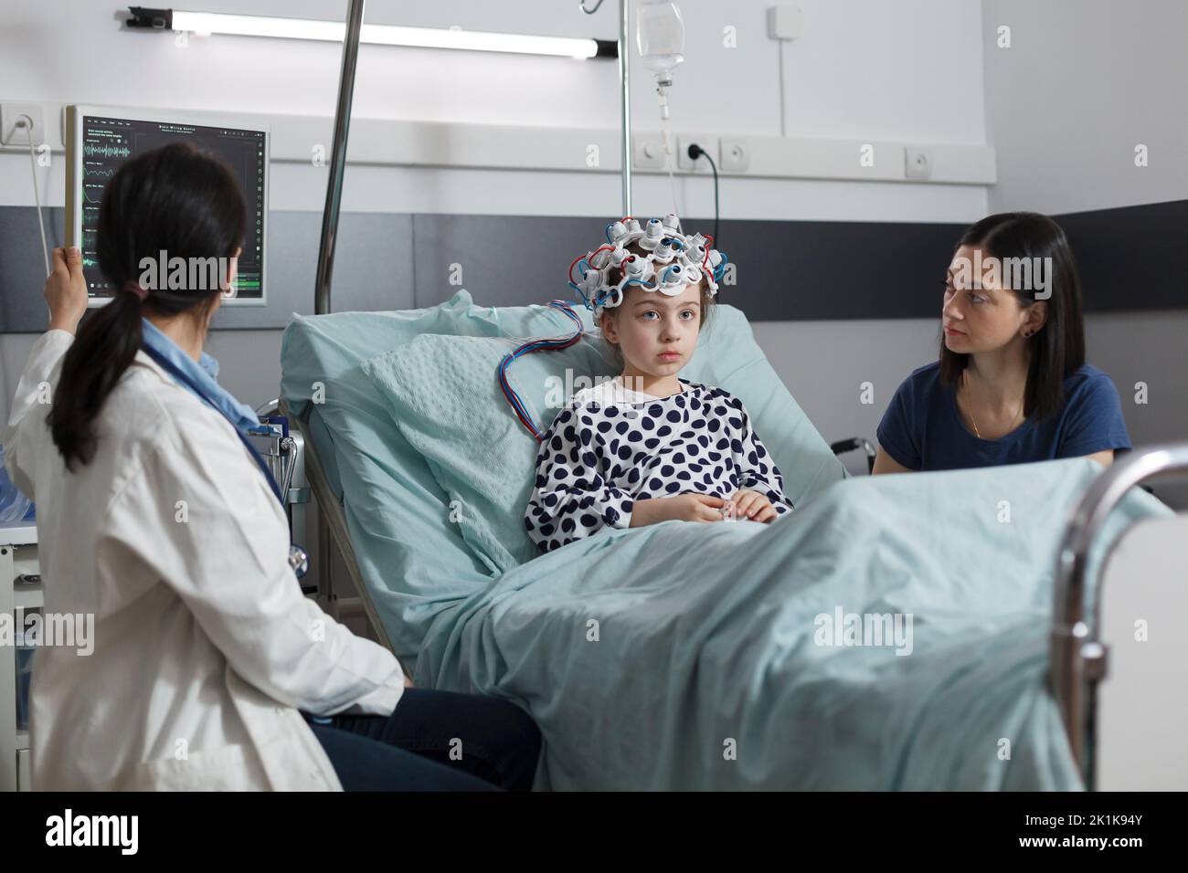 Un pédiatre expert surveille l'état du cerveau des filles hospitalisées à l'aide d'un casque EEG. Spécialiste en neurologie mesurant l'évolution de la maladie neurologique d'un enfant malade assis dans un établissement de soins pédiatriques. Banque D'Images