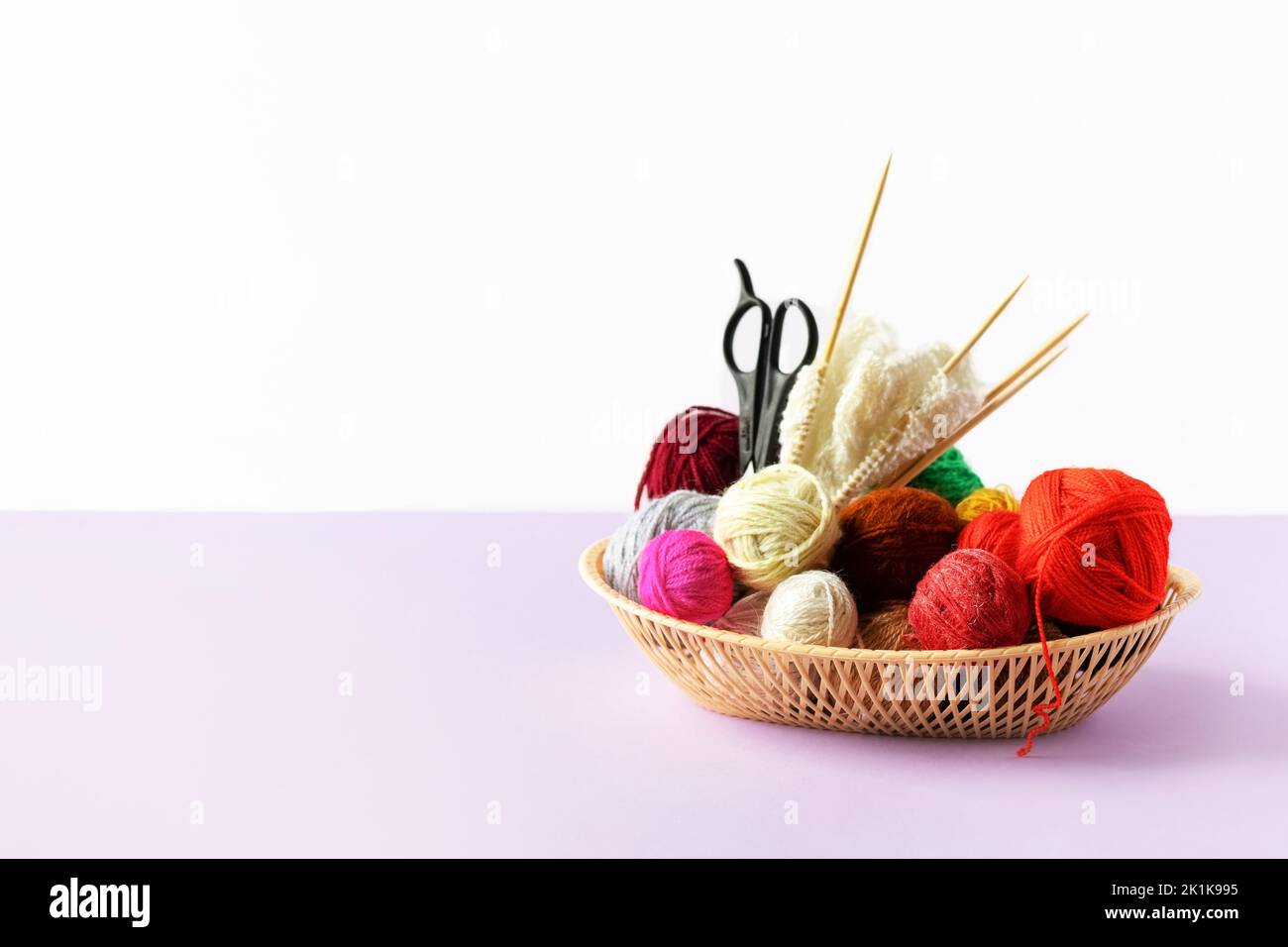 Rouge, jaune, blanc, violet, marron, boules vertes de fil dans un panier beige avec aiguilles à tricoter en bambou et ciseaux sur fond lilas Banque D'Images