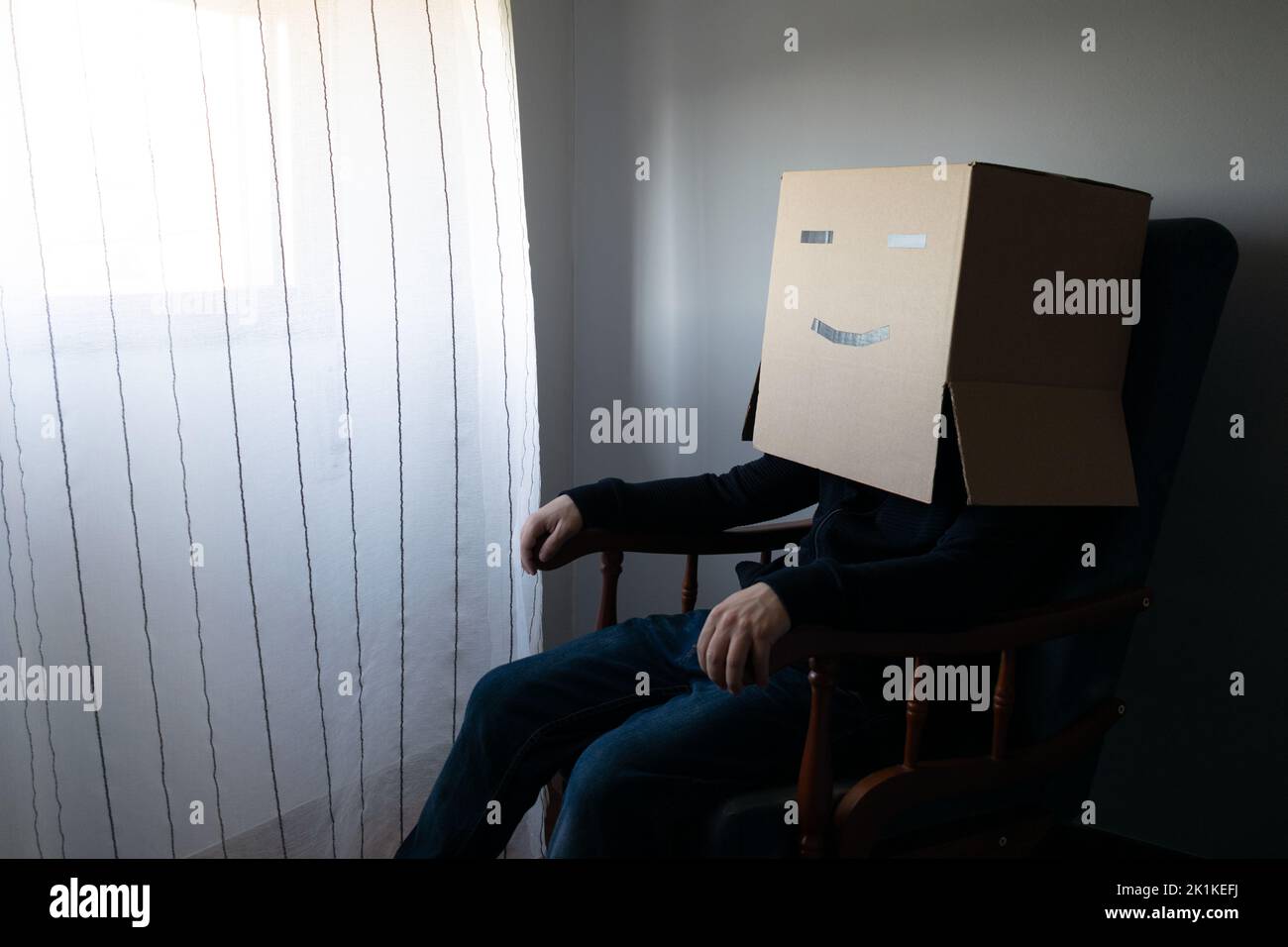 Homme avec une boîte en carton sur sa tête assis sur une chaise en regardant par la fenêtre Banque D'Images