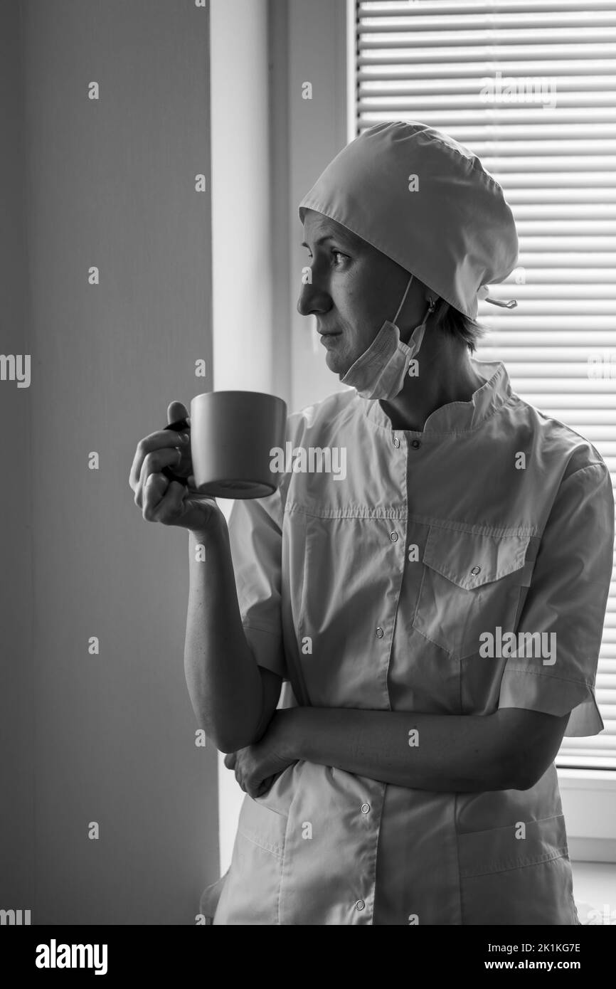 Une femme médecin se tient à la fenêtre de l'hôpital buvant du café. Photo en noir et blanc. Banque D'Images