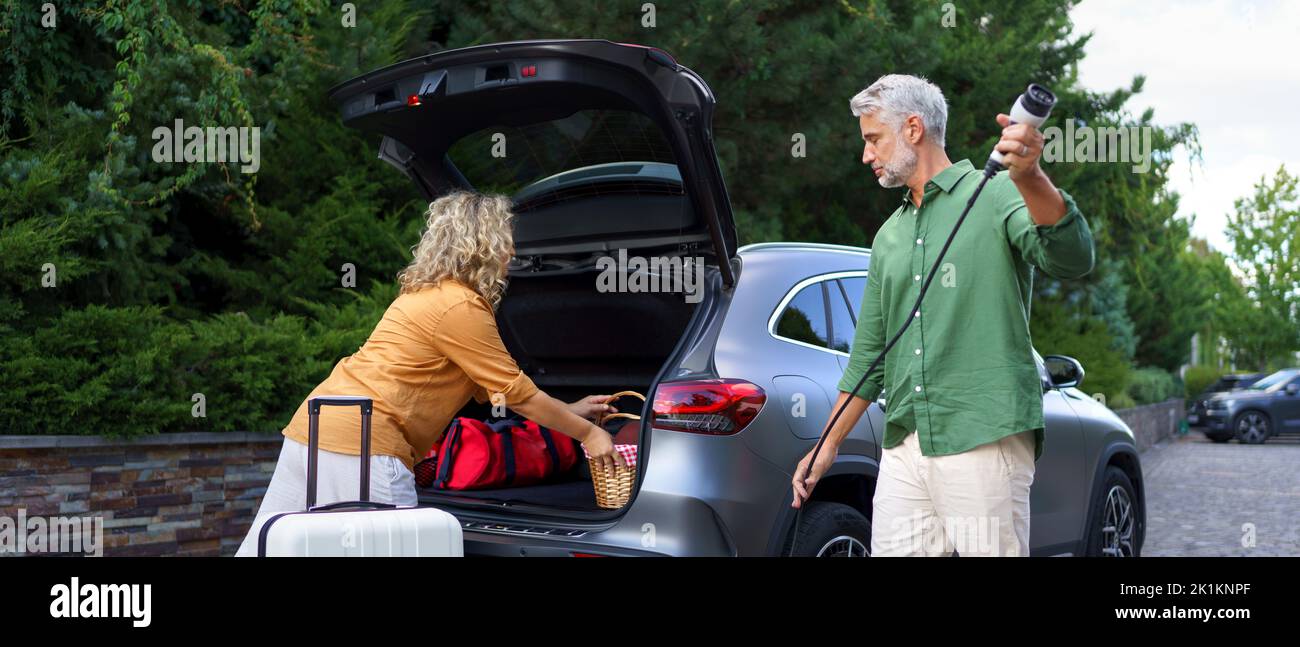 Homme tenant le câble d'alimentation, tandis que sa femme attendant de charger la voiture à la station de charge de véhicule électrique, près Banque D'Images