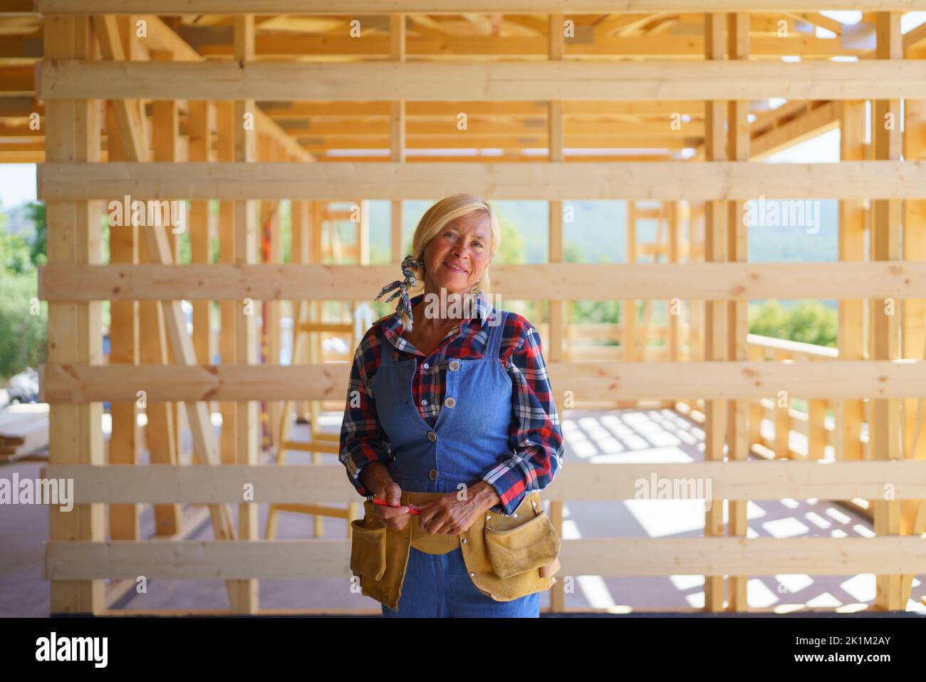 Femme âgée en vêtements de travail posant à l'intérieur de son éco-maison en bois durable écologique inachevé. Concept de femmes et de personnes âgées actives et indépendantes Banque D'Images