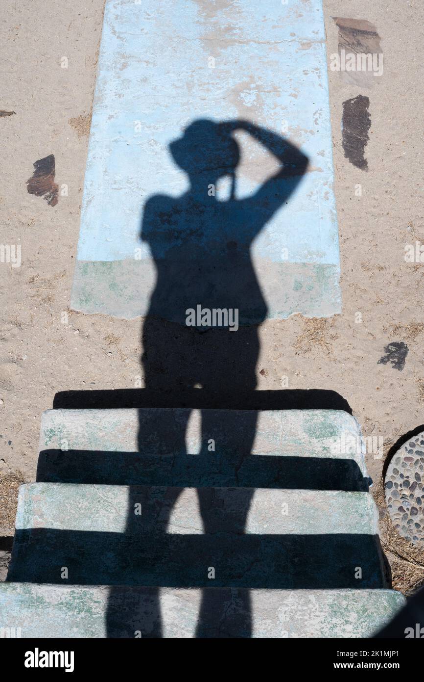 Un photographe avec un chapeau photographie son ombre tombant sur un escalier, du sable et du béton. Banque D'Images