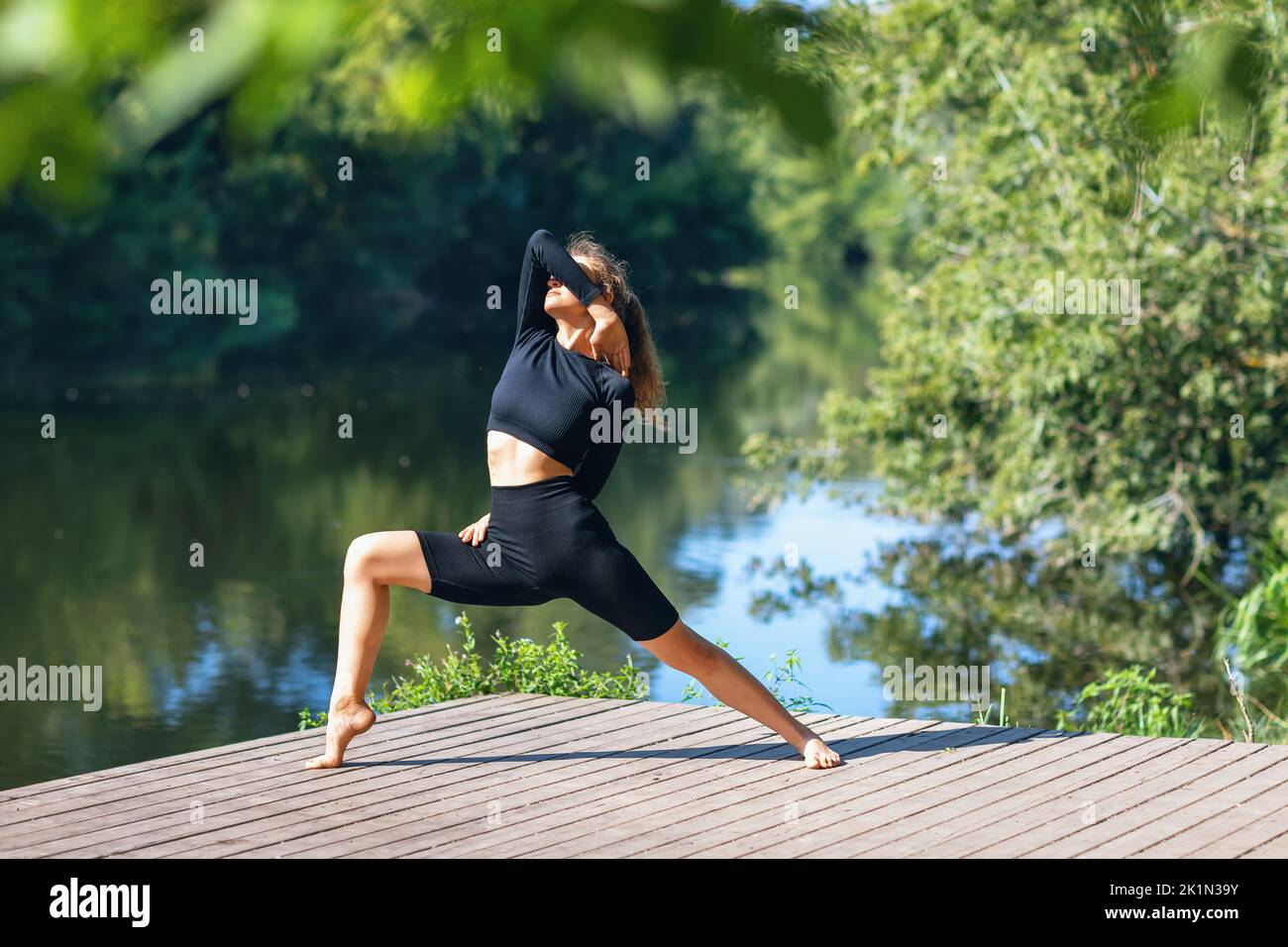 Une femme en vêtements noirs de sport pratiquant le yoga effectue une variation de l'exercice de virabhadrasana, posture de guerrier, s'entraîne sur un chaud matin d'été qui Banque D'Images