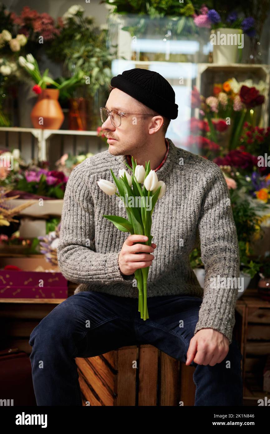 Mignon jeune caesian hipster fleuriste portant des lunettes et beanie en magasin de fleurs avec bouquet de tulipes blanches. Bon propriétaire de magasin de fleurs de sexe masculin dans le chandail. Concept de propriétaire de petite entreprise Banque D'Images