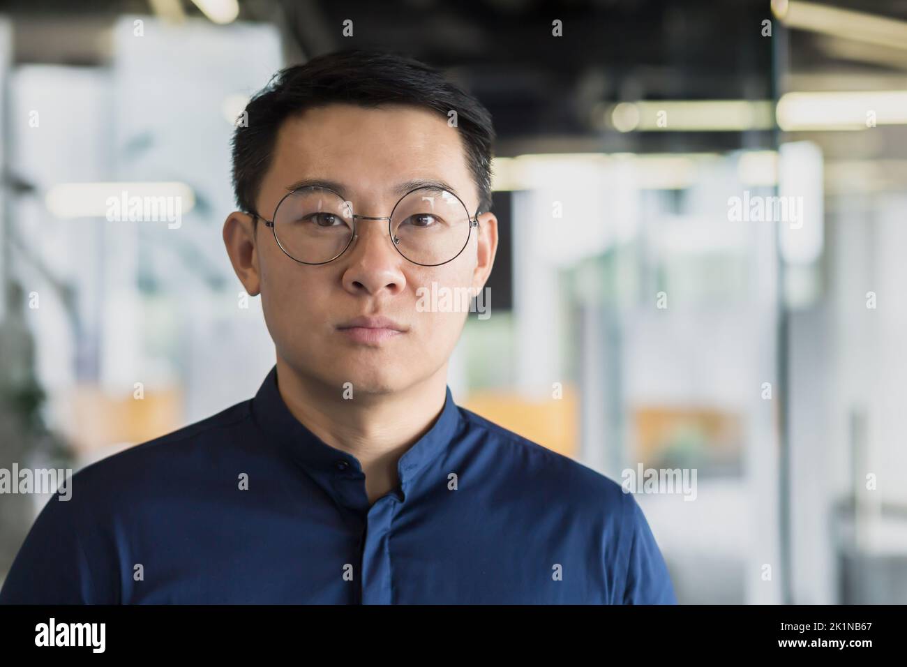 Gros plan portrait d'un investisseur asiatique réussi en lunettes et en chemise bleue, homme d'affaires regardant attentivement l'appareil photo, homme travaillant dans un immeuble de bureaux moderne, patron expérimenté et mature. Banque D'Images