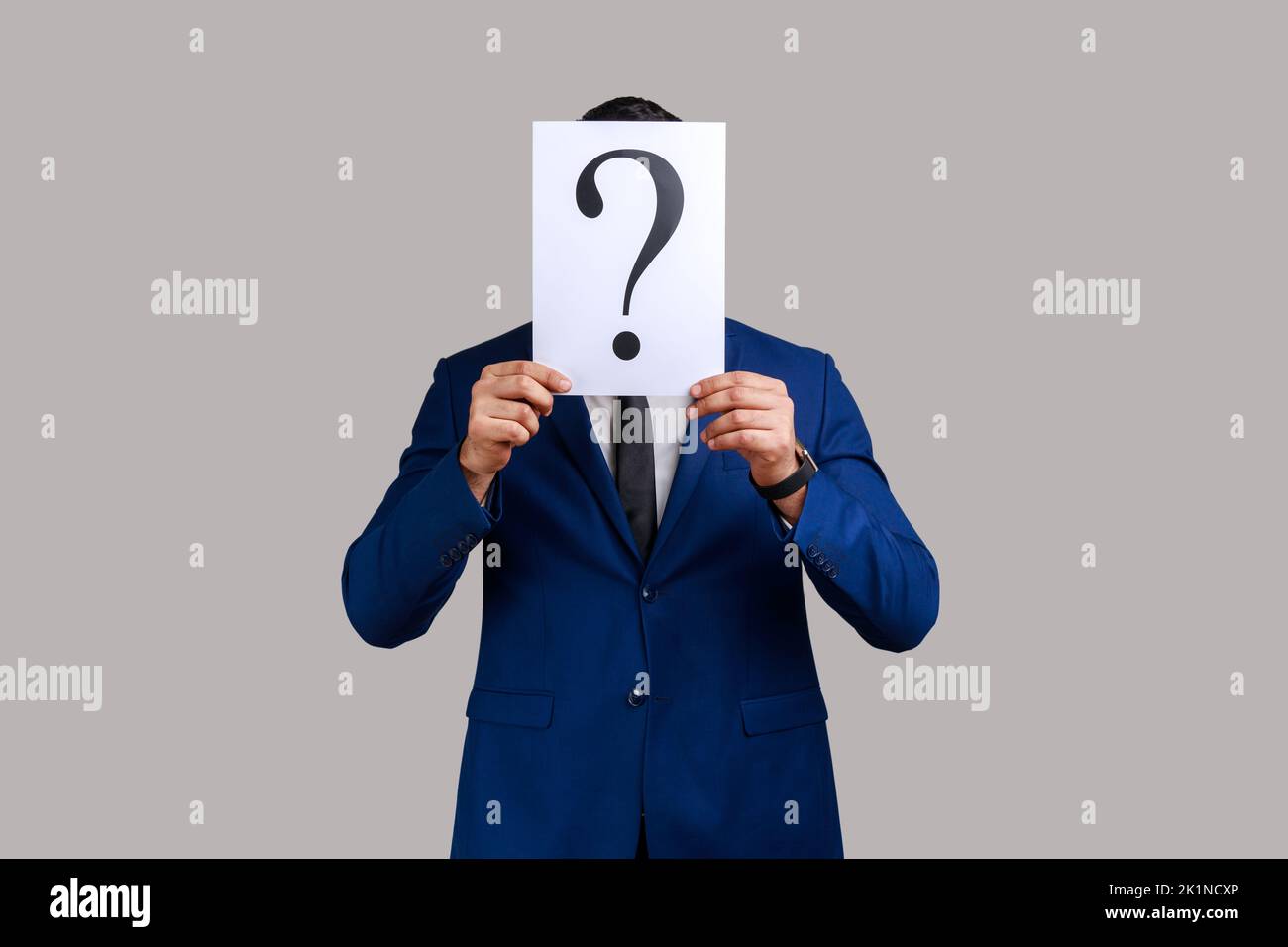Un inconnu qui cache son visage derrière un livre blanc avec un point d'interrogation, trouve une solution intelligente, demande des conseils, porte un costume officiel. Prise de vue en studio isolée sur fond gris. Banque D'Images