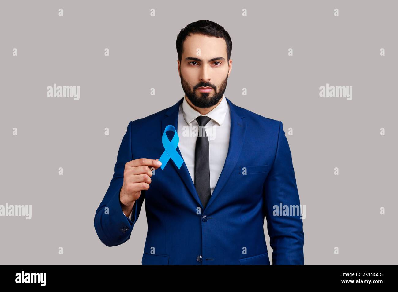 Sensibilisation cancer de la prostate santé des hommes. Portrait d'un homme barbu sérieux tenant le ruban bleu dans les mains, portant un costume de style officiel. Prise de vue en studio isolée sur fond gris. Banque D'Images