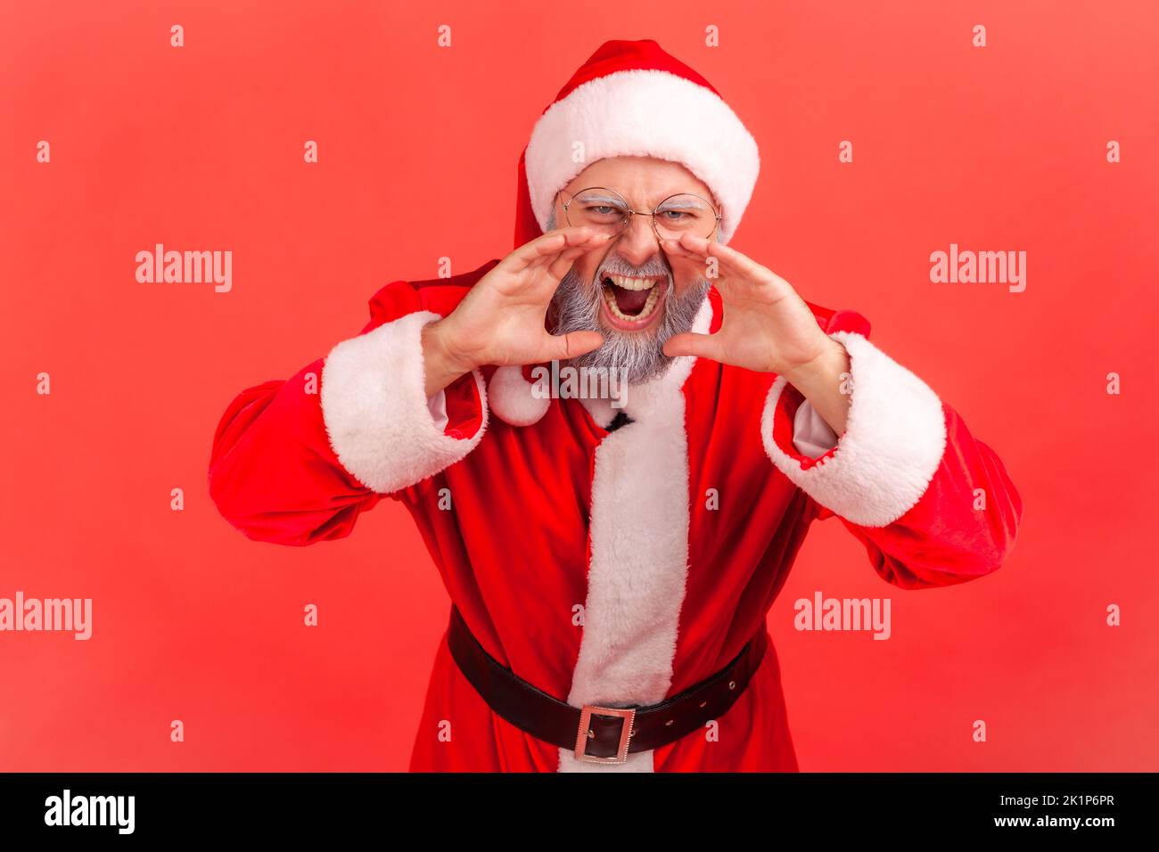 Portrait d'un homme âgé avec une barbe grise portant le costume du père noël criant avec une expression faciale en colère, en gardant les mains près de la bouche. Studio d'intérieur isolé sur fond rouge. Banque D'Images