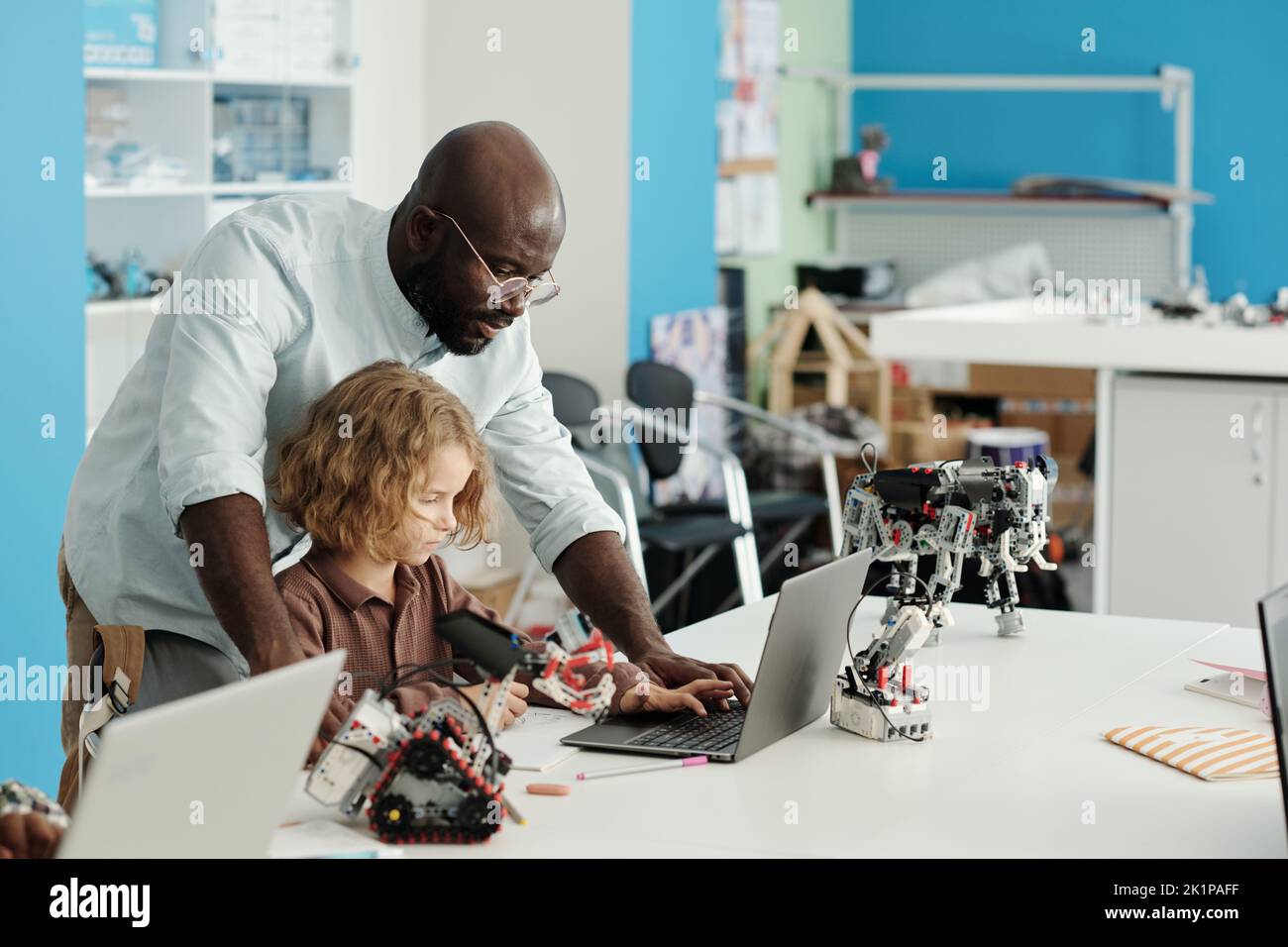 Un jeune professeur afro-américain de robotique se penche sur un adorable écolier assis près d'un bureau devant un ordinateur portable et travaillant sur un nouveau projet Banque D'Images